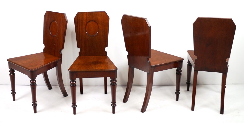 A set of four Victorian mahogany hall chairs featuring shaped solid wooden backs with recessed circular central details, flat wooden seats, turned front legs, and flared rear legs. View III