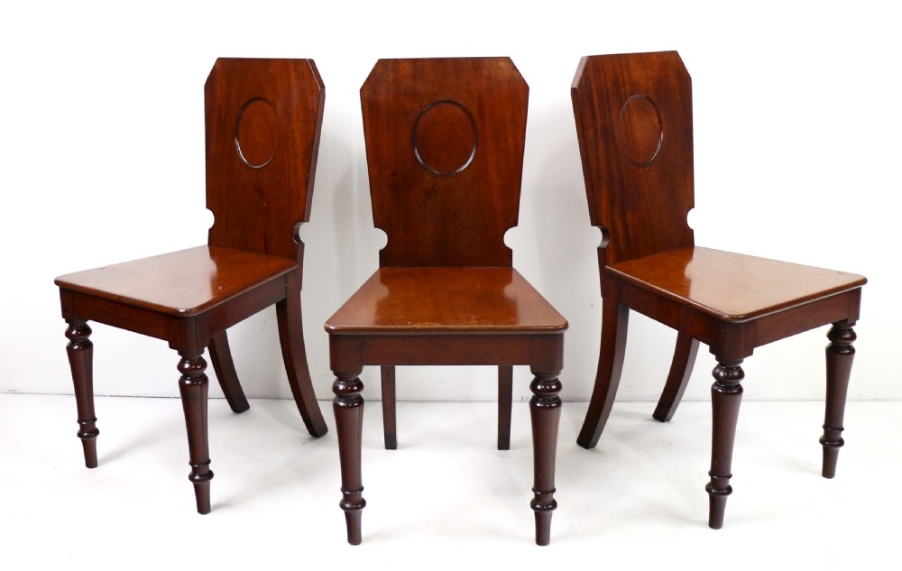 A set of four Victorian mahogany hall chairs featuring shaped solid wooden backs with recessed circular central details, flat wooden seats, turned front legs, and flared rear legs. View V