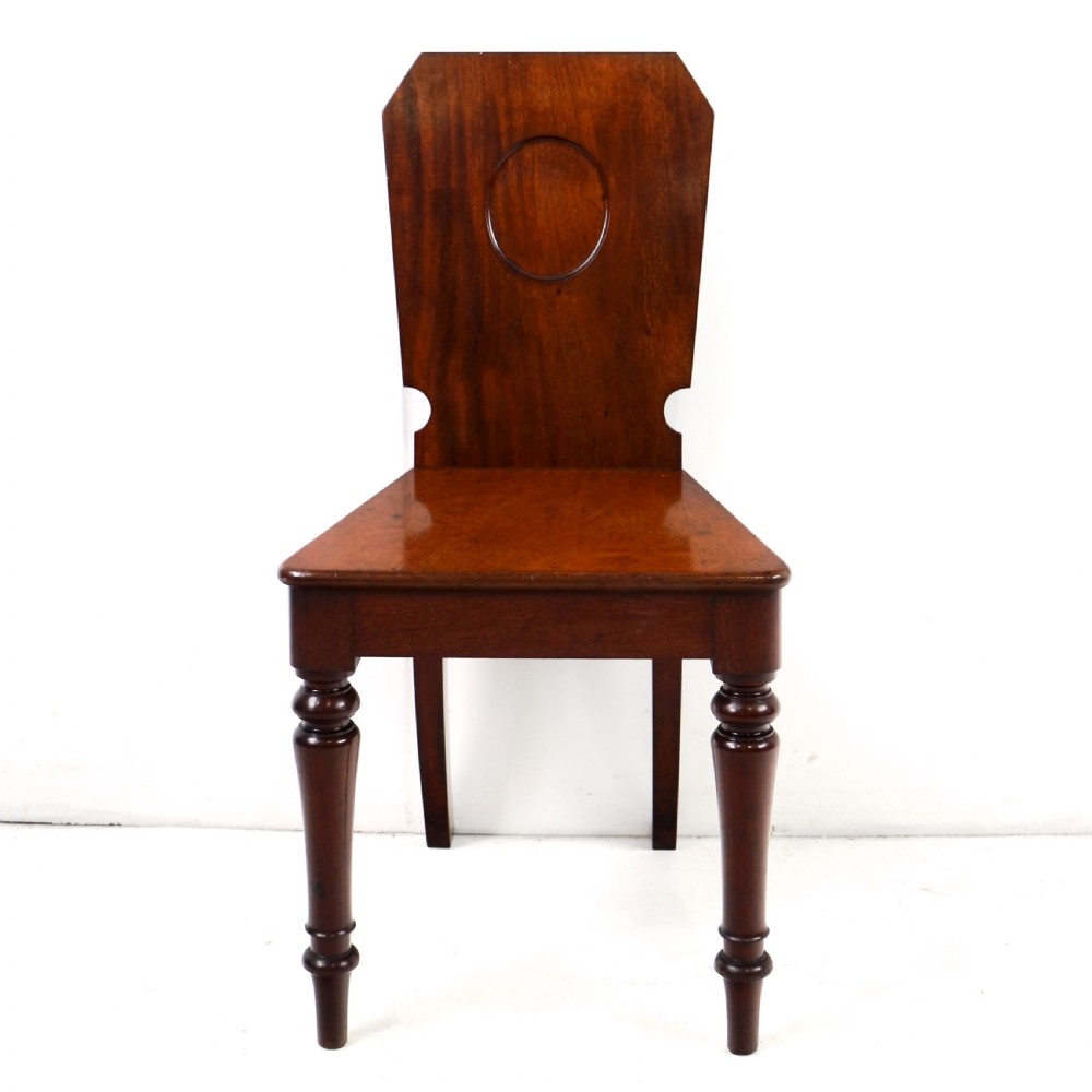 A set of four Victorian mahogany hall chairs featuring shaped solid wooden backs with recessed circular central details, flat wooden seats, turned front legs, and flared rear legs. View VI