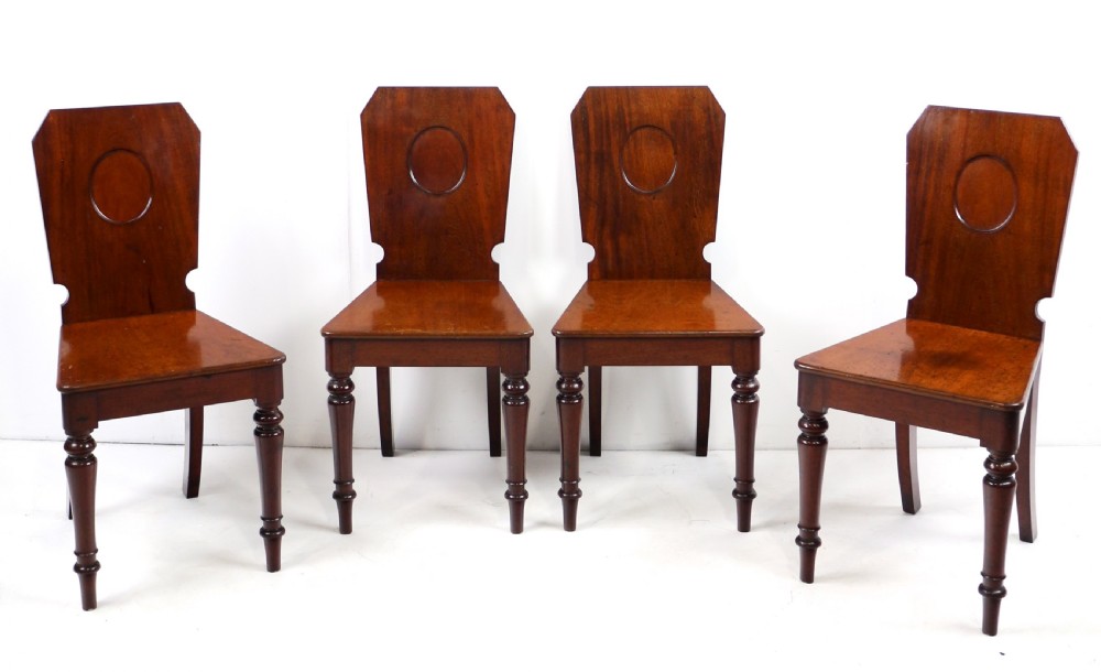 A set of four Victorian mahogany hall chairs featuring shaped solid wooden backs with recessed circular central details, flat wooden seats, turned front legs, and flared rear legs. View VII