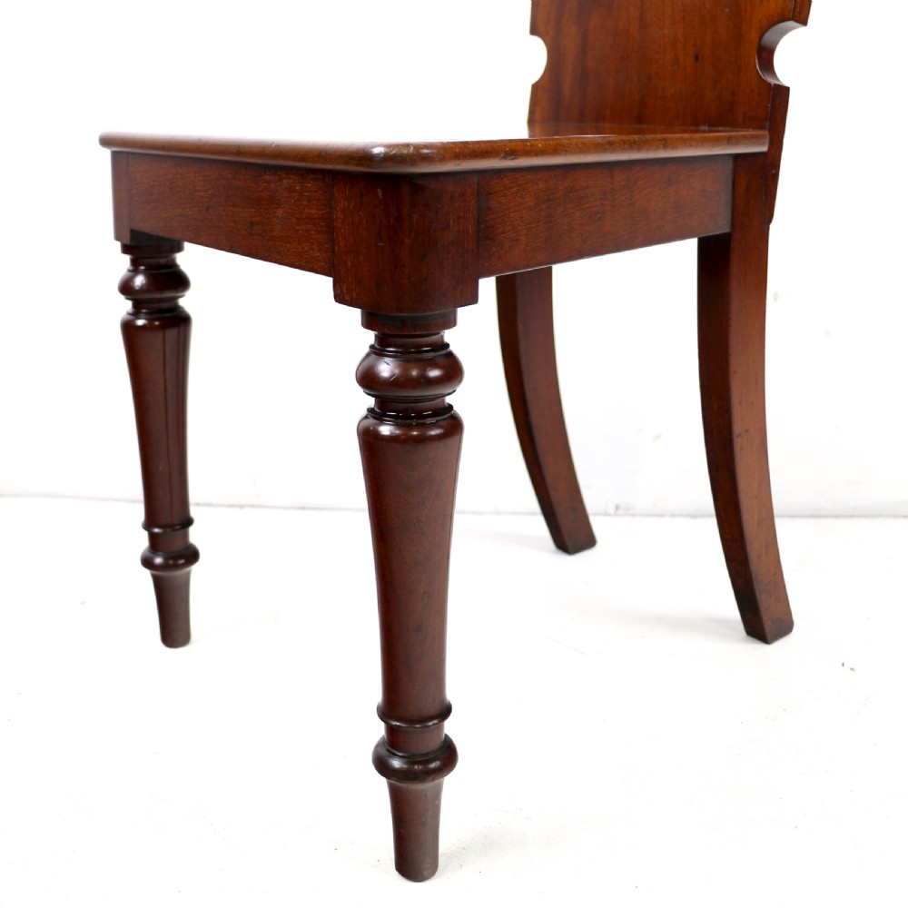 A set of four Victorian mahogany hall chairs featuring shaped solid wooden backs with recessed circular central details, flat wooden seats, turned front legs, and flared rear legs. View X