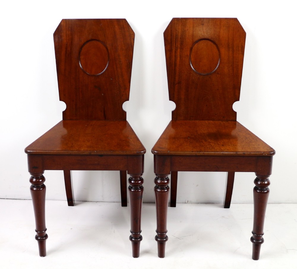 A set of four Victorian mahogany hall chairs featuring shaped solid wooden backs with recessed circular central details, flat wooden seats, turned front legs, and flared rear legs. View XIII