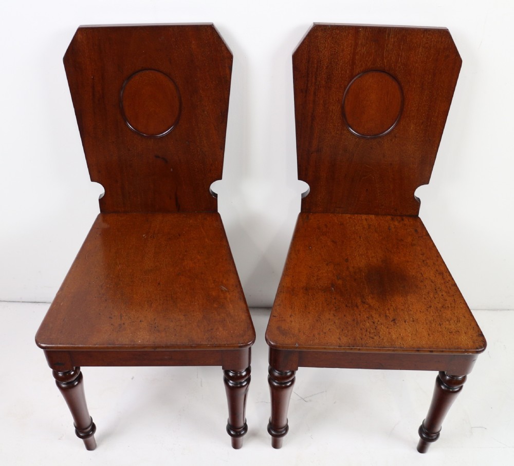 A set of four Victorian mahogany hall chairs featuring shaped solid wooden backs with recessed circular central details, flat wooden seats, turned front legs, and flared rear legs. View XIV