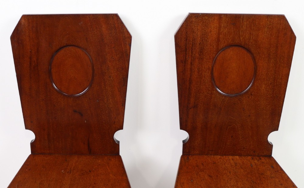 A set of four Victorian mahogany hall chairs featuring shaped solid wooden backs with recessed circular central details, flat wooden seats, turned front legs, and flared rear legs. View XV