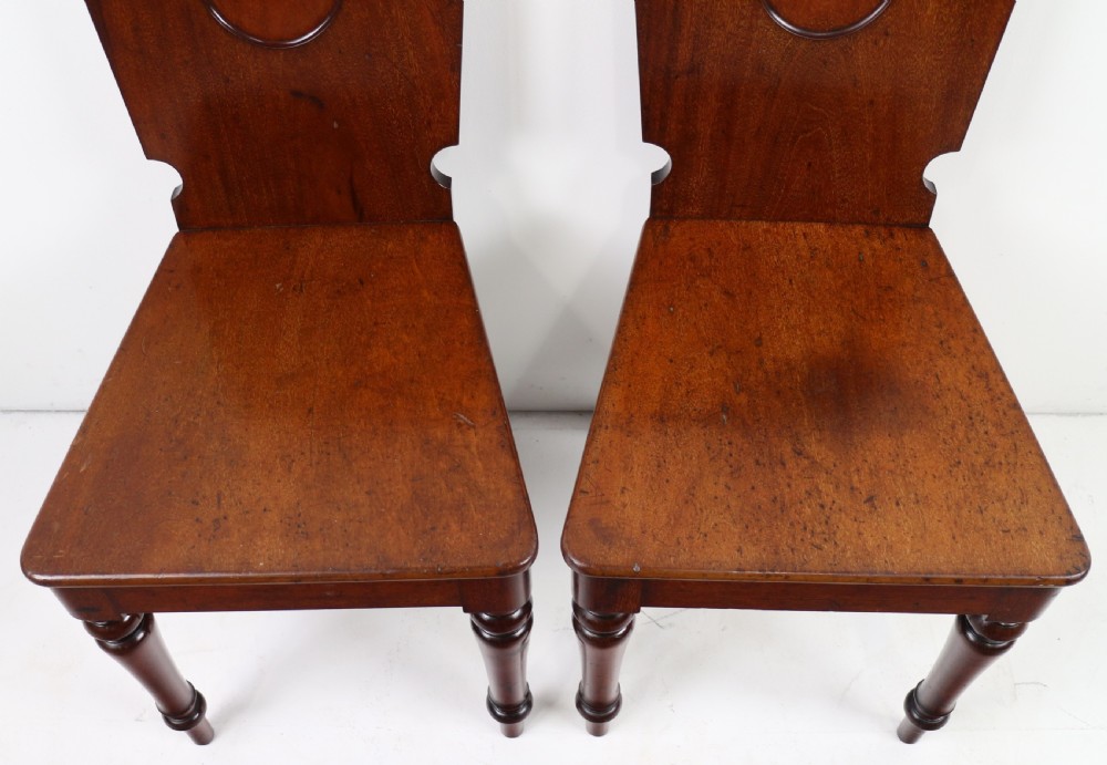 A set of four Victorian mahogany hall chairs featuring shaped solid wooden backs with recessed circular central details, flat wooden seats, turned front legs, and flared rear legs. View XVI