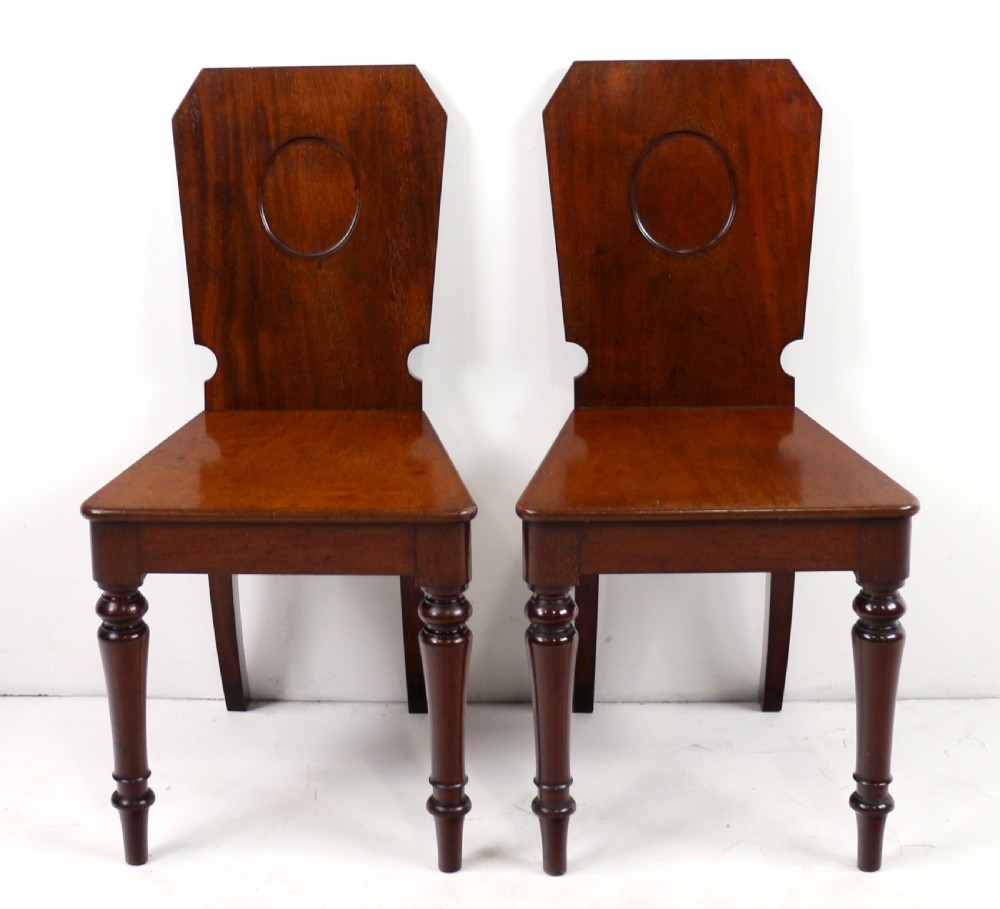 A set of four Victorian mahogany hall chairs featuring shaped solid wooden backs with recessed circular central details, flat wooden seats, turned front legs, and flared rear legs. View XVIII