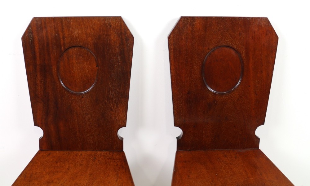 A set of four Victorian mahogany hall chairs featuring shaped solid wooden backs with recessed circular central details, flat wooden seats, turned front legs, and flared rear legs. View XIX