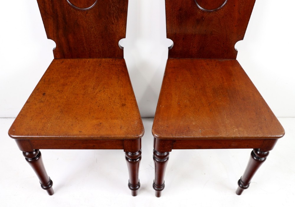 A set of four Victorian mahogany hall chairs featuring shaped solid wooden backs with recessed circular central details, flat wooden seats, turned front legs, and flared rear legs. View XX