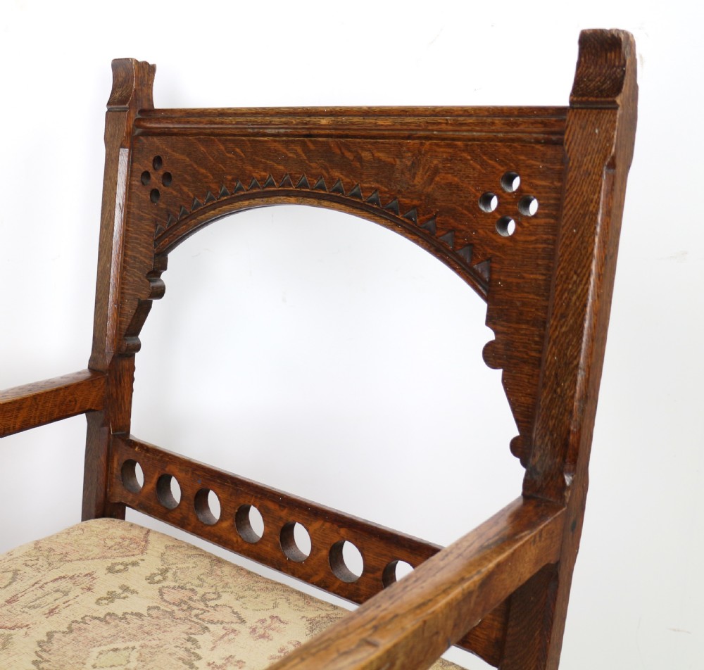 A Gothic Revival carved dark oak armchair featuring an arched backrest with quatrefoil cutouts, curved legs, and a patterned fabric seat. View X