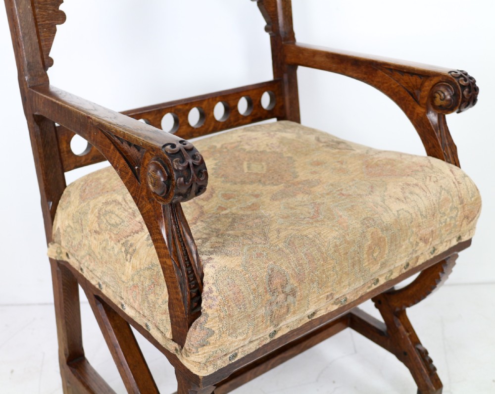 A Gothic Revival carved dark oak armchair featuring an arched backrest with quatrefoil cutouts, curved legs, and a patterned fabric seat. View XII