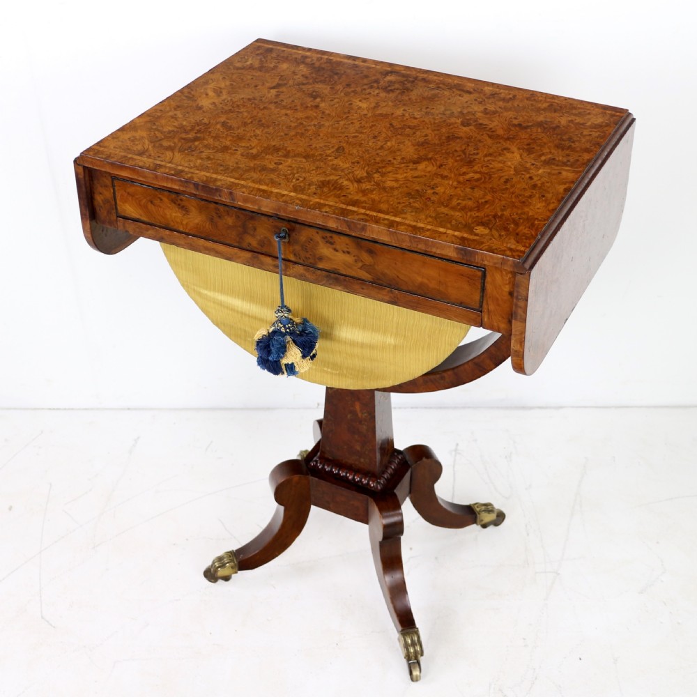 An Irish Regency drop-leaf work table featuring burr yew veneer, a single drawer, a yellow pleated fabric work well, and a quadruped pedestal base with brass castors.