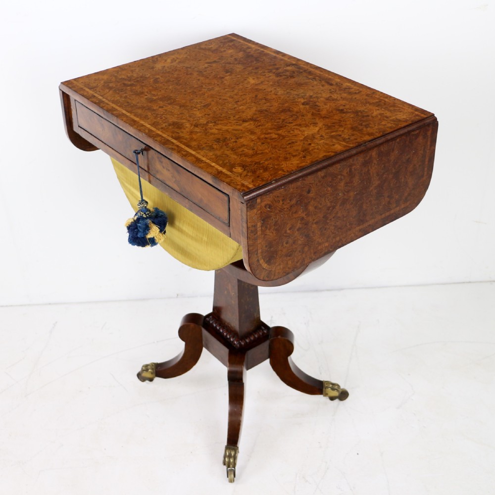 An Irish Regency drop-leaf work table featuring burr yew veneer, a single drawer, a yellow pleated fabric work well, and a quadruped pedestal base with brass castors. View V