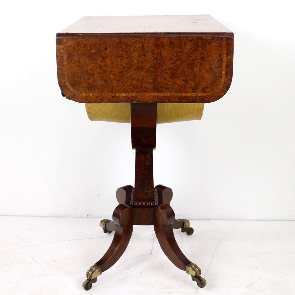 An Irish Regency drop-leaf work table featuring burr yew veneer, a single drawer, a yellow pleated fabric work well, and a quadruped pedestal base with brass castors. View X