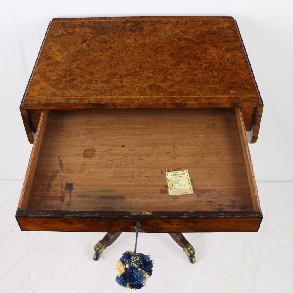 An Irish Regency drop-leaf work table featuring burr yew veneer, a single drawer, a yellow pleated fabric work well, and a quadruped pedestal base with brass castors. View XVII