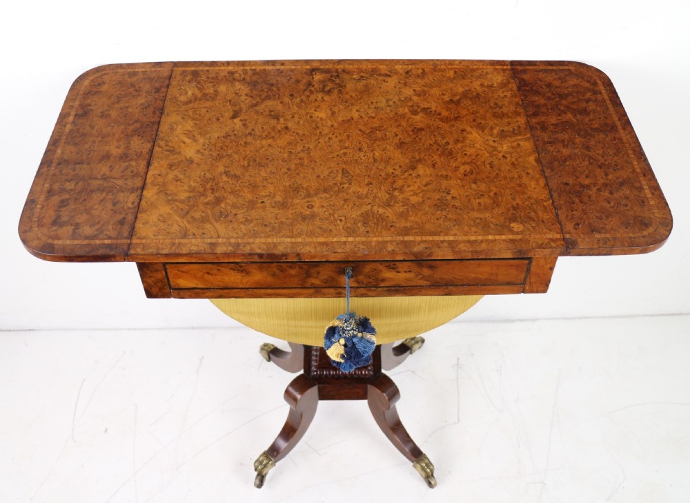 An Irish Regency drop-leaf work table featuring burr yew veneer, a single drawer, a yellow pleated fabric work well, and a quadruped pedestal base with brass castors. View XXVI