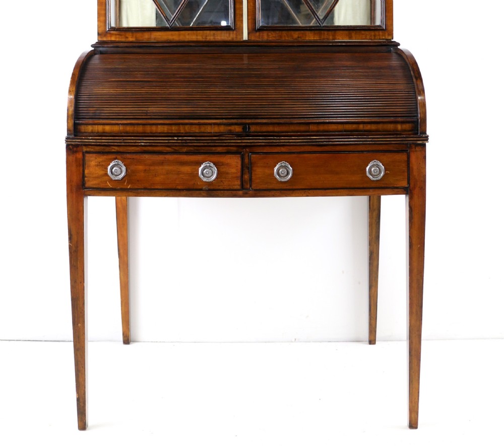 An antique mahogany writing desk cabinet featuring an upper glass cabinet with diamond glazing bars, a curved slatted tambour shutter section, and two drawers on square tapered legs. View VI