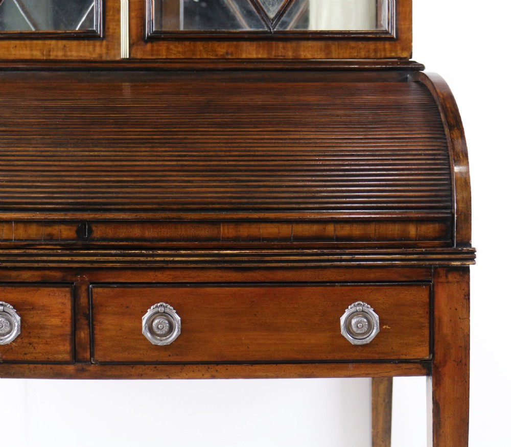 An antique mahogany writing desk cabinet featuring an upper glass cabinet with diamond glazing bars, a curved slatted tambour shutter section, and two drawers on square tapered legs. View VII