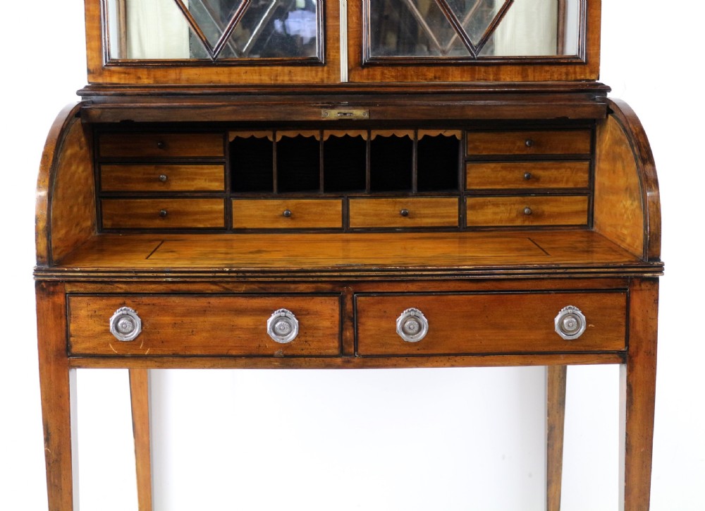 An antique mahogany writing desk cabinet featuring an upper glass cabinet with diamond glazing bars, a curved slatted tambour shutter section, and two drawers on square tapered legs. View XXI