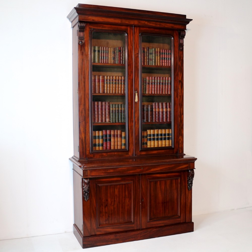 A tall William IV Cuban mahogany bookcase featuring a glazed two-door upper section with shelving above a two-door panelled cupboard base with carved side corbels. View II