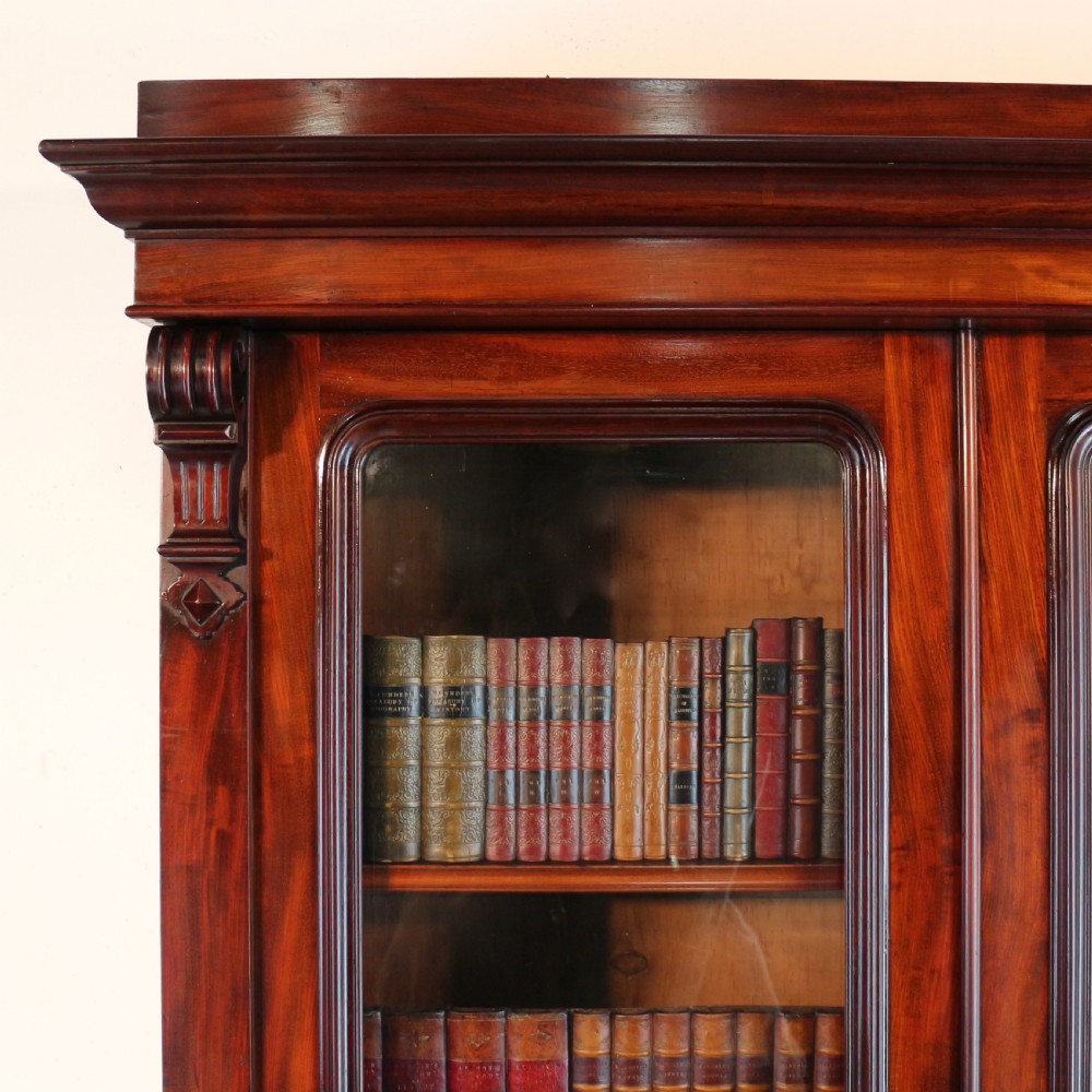 A tall William IV Cuban mahogany bookcase featuring a glazed two-door upper section with shelving above a two-door panelled cupboard base with carved side corbels. View V