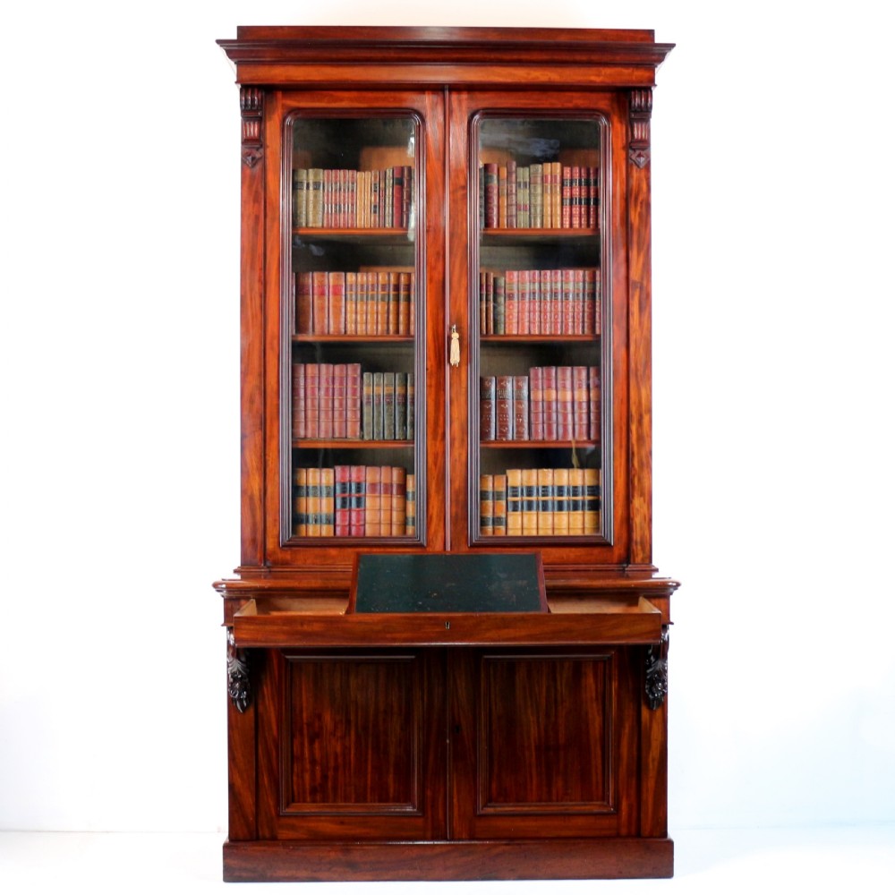 A tall William IV Cuban mahogany bookcase featuring a glazed two-door upper section with shelving above a two-door panelled cupboard base with carved side corbels. View VI