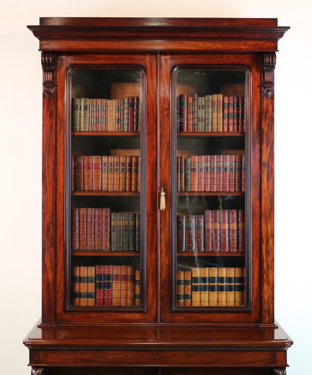 A tall William IV Cuban mahogany bookcase featuring a glazed two-door upper section with shelving above a two-door panelled cupboard base with carved side corbels. View VII