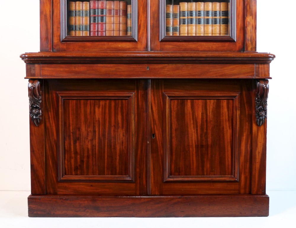 A tall William IV Cuban mahogany bookcase featuring a glazed two-door upper section with shelving above a two-door panelled cupboard base with carved side corbels. View VIII