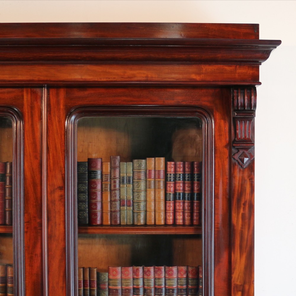 A tall William IV Cuban mahogany bookcase featuring a glazed two-door upper section with shelving above a two-door panelled cupboard base with carved side corbels. View IX