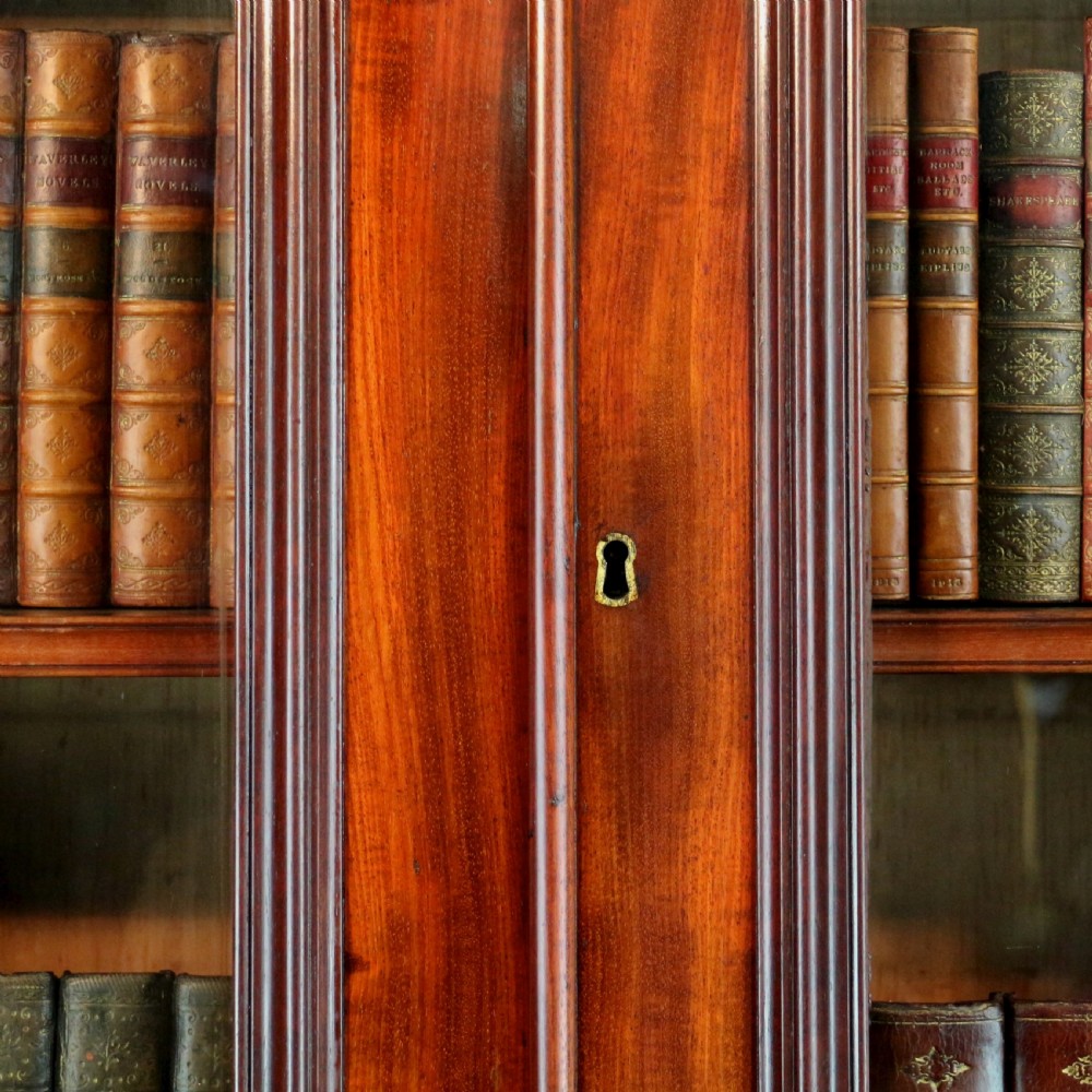A tall William IV Cuban mahogany bookcase featuring a glazed two-door upper section with shelving above a two-door panelled cupboard base with carved side corbels. View XI