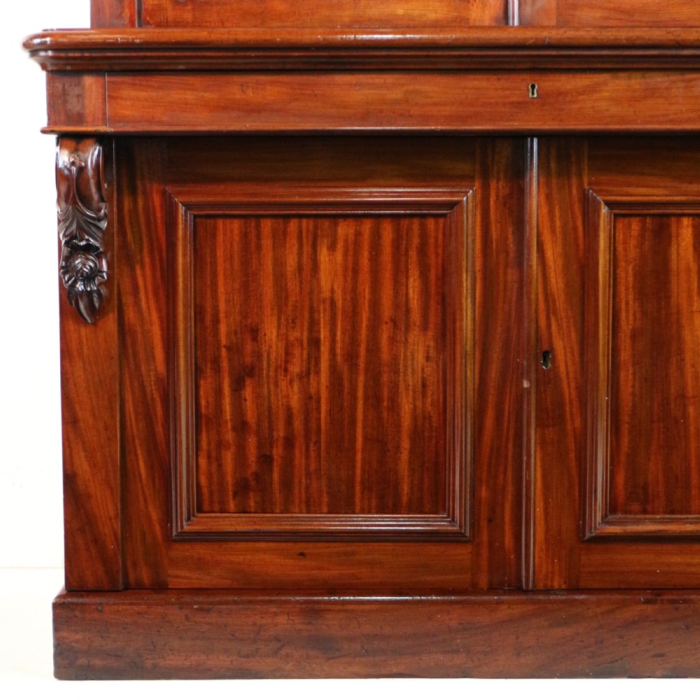 A tall William IV Cuban mahogany bookcase featuring a glazed two-door upper section with shelving above a two-door panelled cupboard base with carved side corbels. View XII