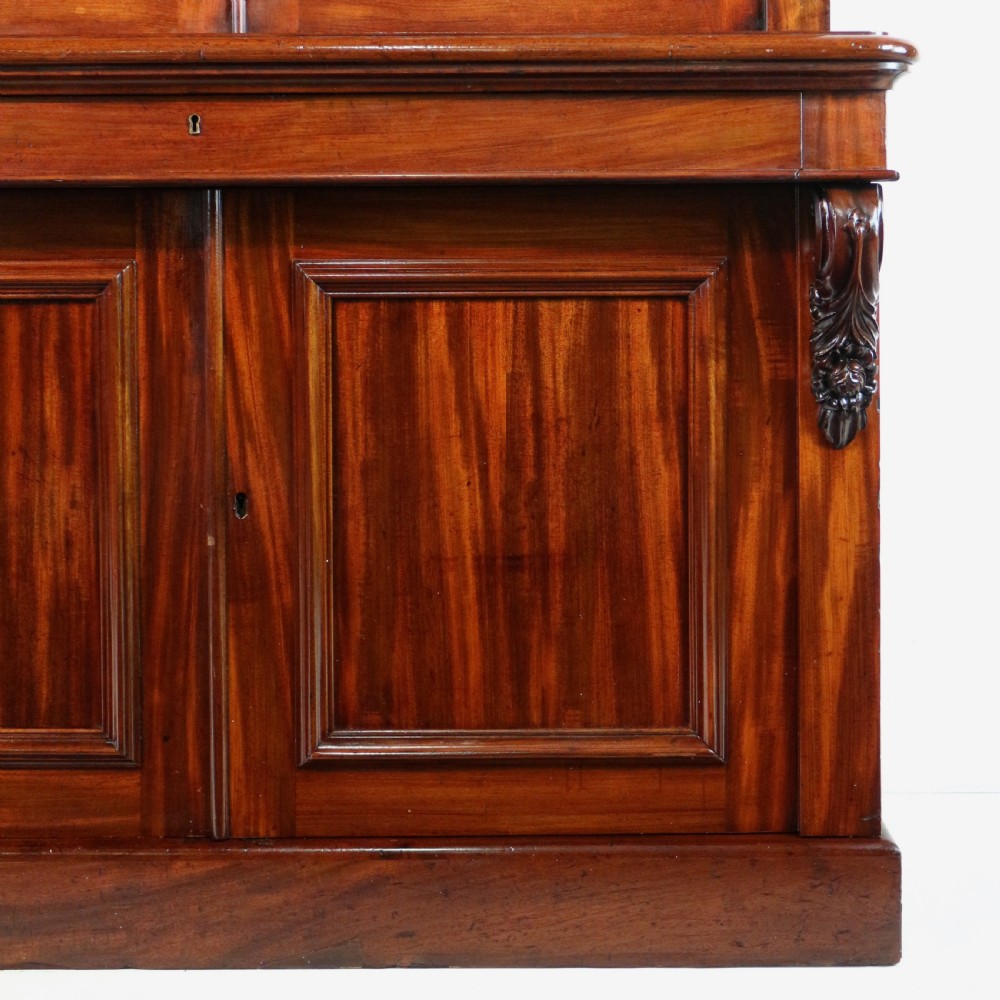 A tall William IV Cuban mahogany bookcase featuring a glazed two-door upper section with shelving above a two-door panelled cupboard base with carved side corbels. View XIII