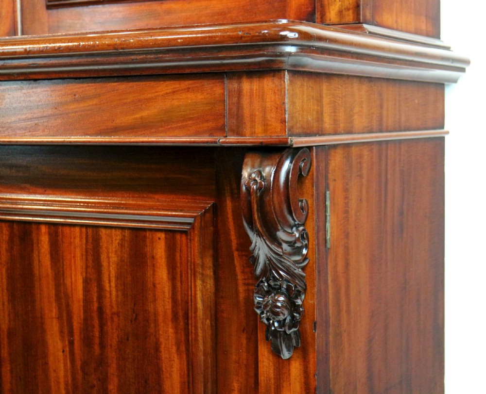 A tall William IV Cuban mahogany bookcase featuring a glazed two-door upper section with shelving above a two-door panelled cupboard base with carved side corbels. View XIV