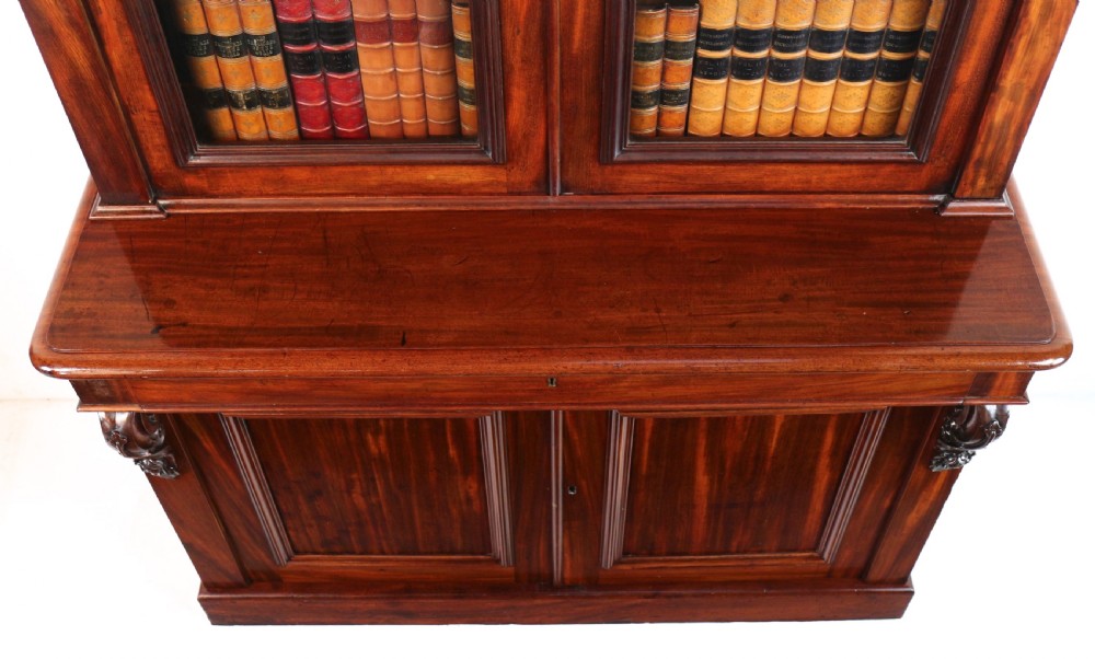 A tall William IV Cuban mahogany bookcase featuring a glazed two-door upper section with shelving above a two-door panelled cupboard base with carved side corbels. View XV