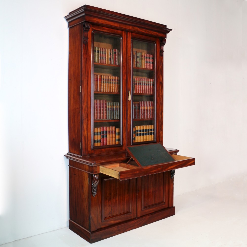 A tall William IV Cuban mahogany bookcase featuring a glazed two-door upper section with shelving above a two-door panelled cupboard base with carved side corbels. View XVI
