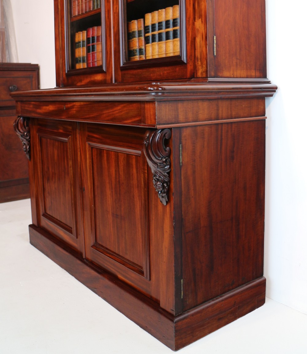 A tall William IV Cuban mahogany bookcase featuring a glazed two-door upper section with shelving above a two-door panelled cupboard base with carved side corbels. View XXIV