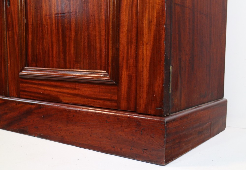 A tall William IV Cuban mahogany bookcase featuring a glazed two-door upper section with shelving above a two-door panelled cupboard base with carved side corbels. View XXV
