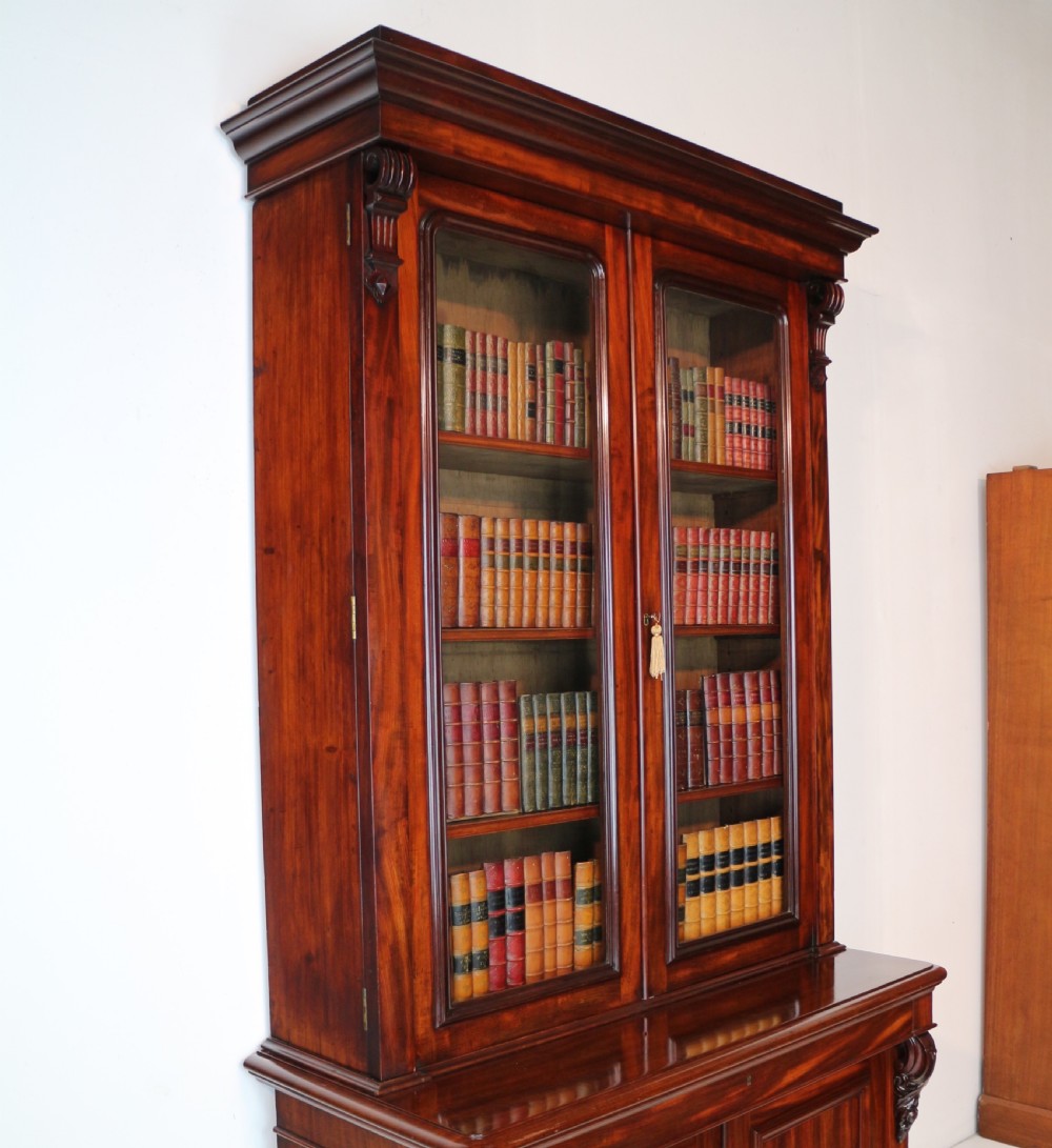 A tall William IV Cuban mahogany bookcase featuring a glazed two-door upper section with shelving above a two-door panelled cupboard base with carved side corbels. View XXVI