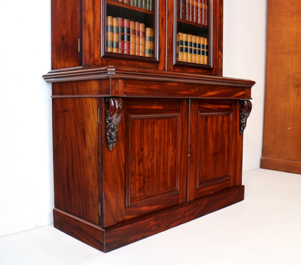 A tall William IV Cuban mahogany bookcase featuring a glazed two-door upper section with shelving above a two-door panelled cupboard base with carved side corbels. View XXVIII