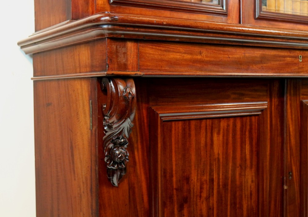 A tall William IV Cuban mahogany bookcase featuring a glazed two-door upper section with shelving above a two-door panelled cupboard base with carved side corbels. View XXIX
