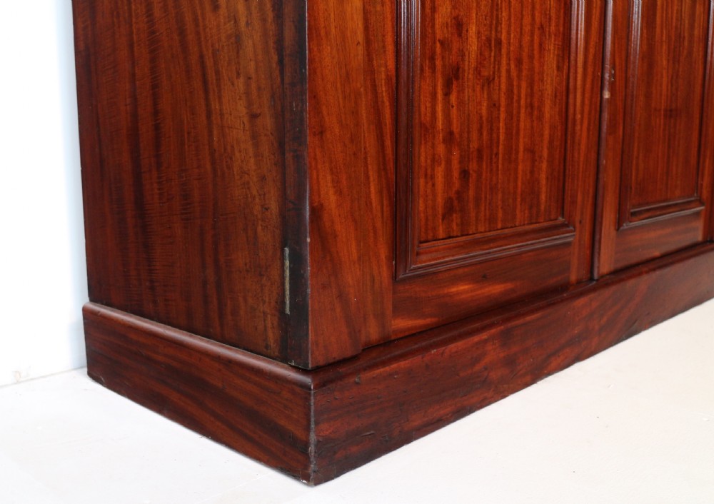 A tall William IV Cuban mahogany bookcase featuring a glazed two-door upper section with shelving above a two-door panelled cupboard base with carved side corbels. View XXX