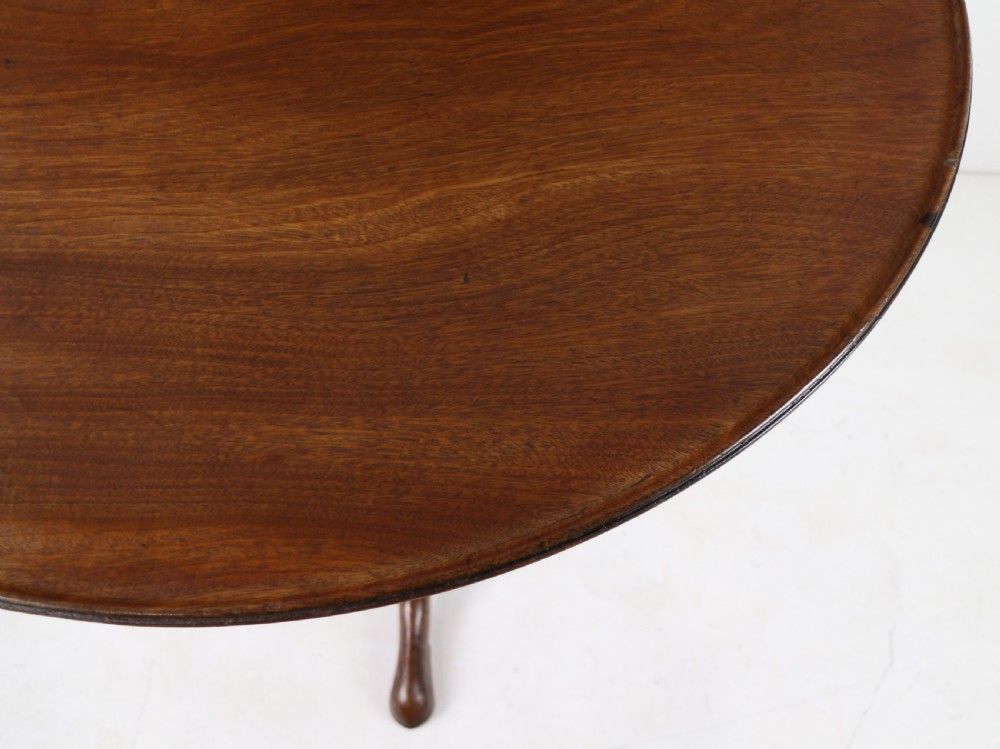 An Early Georgian mahogany tripod table featuring a circular dish top tilted vertically above a turned stem and cabriole legs with pad feet. View XV