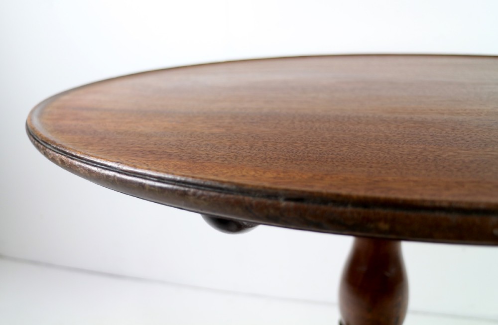 An Early Georgian mahogany tripod table featuring a circular dish top tilted vertically above a turned stem and cabriole legs with pad feet. View XVI