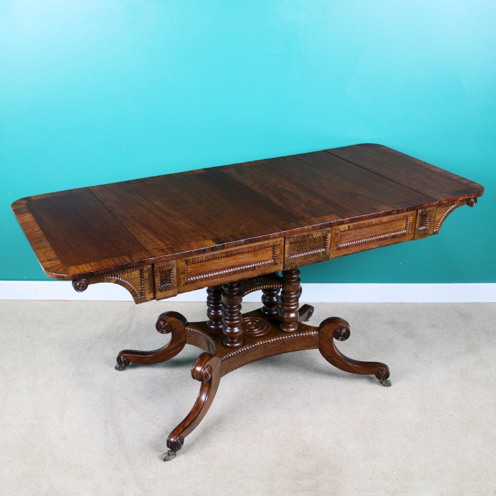 A Regency mahogany and rosewood crossbanded sofa table featuring drop leaves, bead-moulded frieze drawers, turned support pillars, and splayed legs on brass castors.