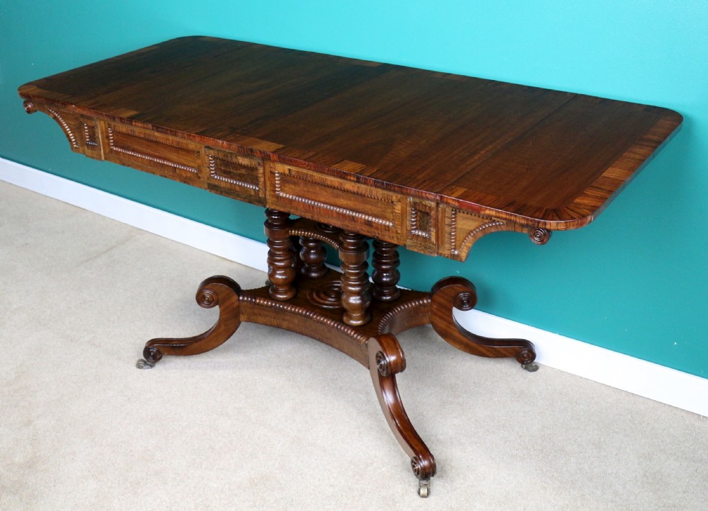 A Regency mahogany and rosewood crossbanded sofa table featuring drop leaves, bead-moulded frieze drawers, turned support pillars, and splayed legs on brass castors. View II