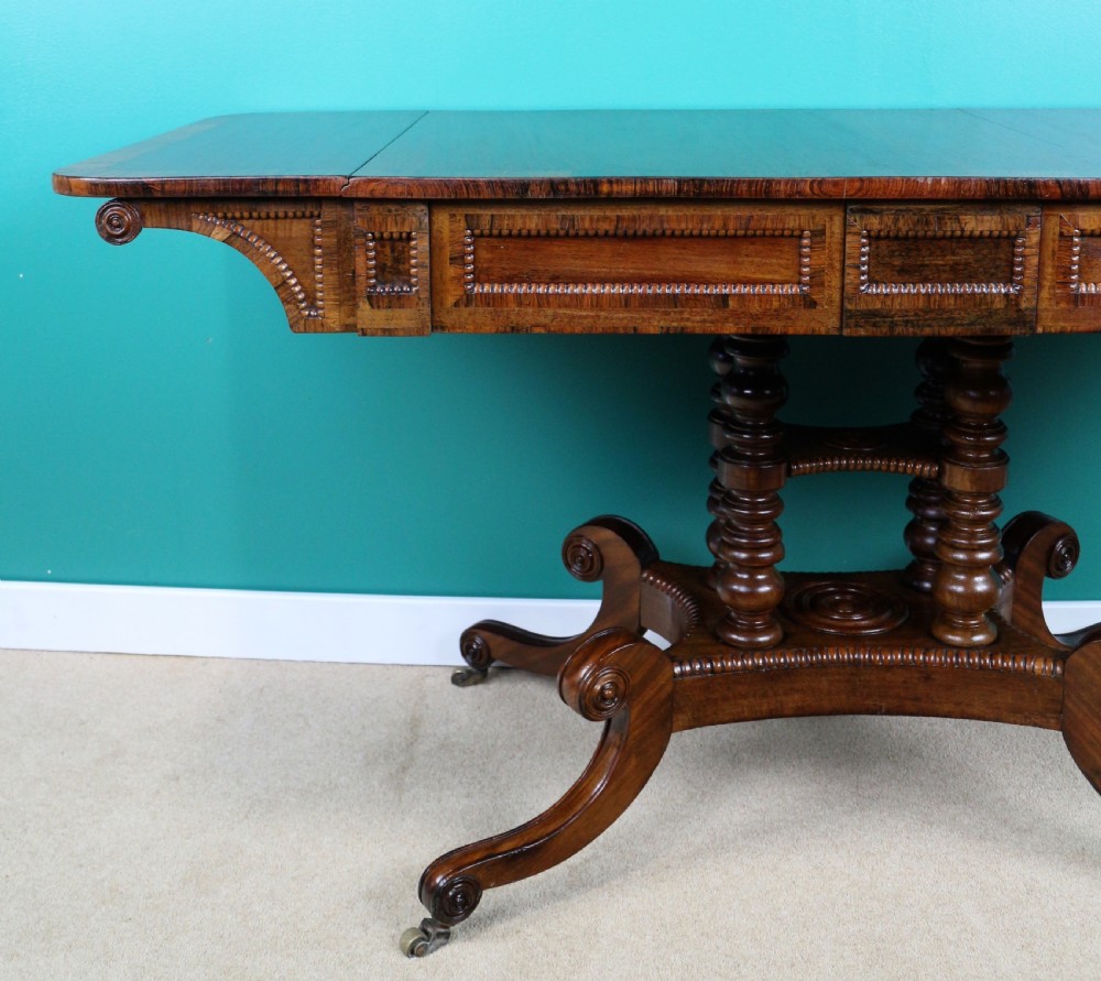 A Regency mahogany and rosewood crossbanded sofa table featuring drop leaves, bead-moulded frieze drawers, turned support pillars, and splayed legs on brass castors. View IV
