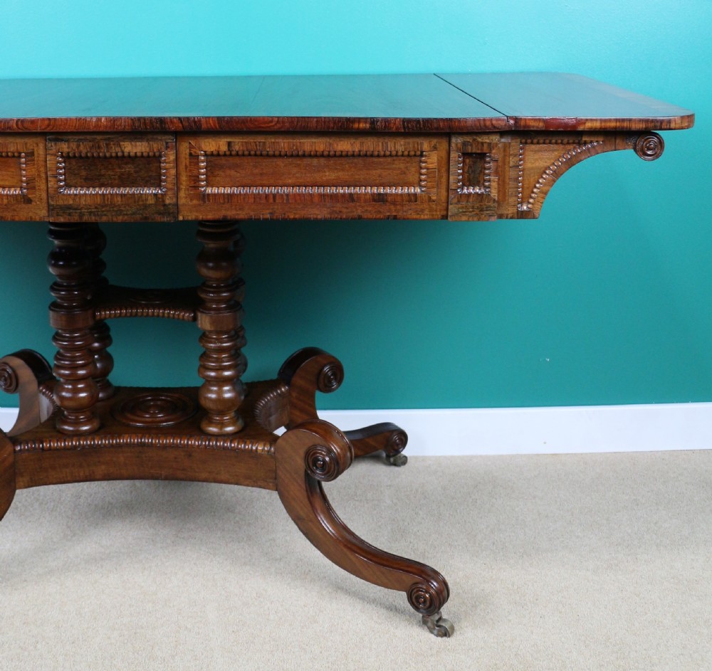 A Regency mahogany and rosewood crossbanded sofa table featuring drop leaves, bead-moulded frieze drawers, turned support pillars, and splayed legs on brass castors. View V
