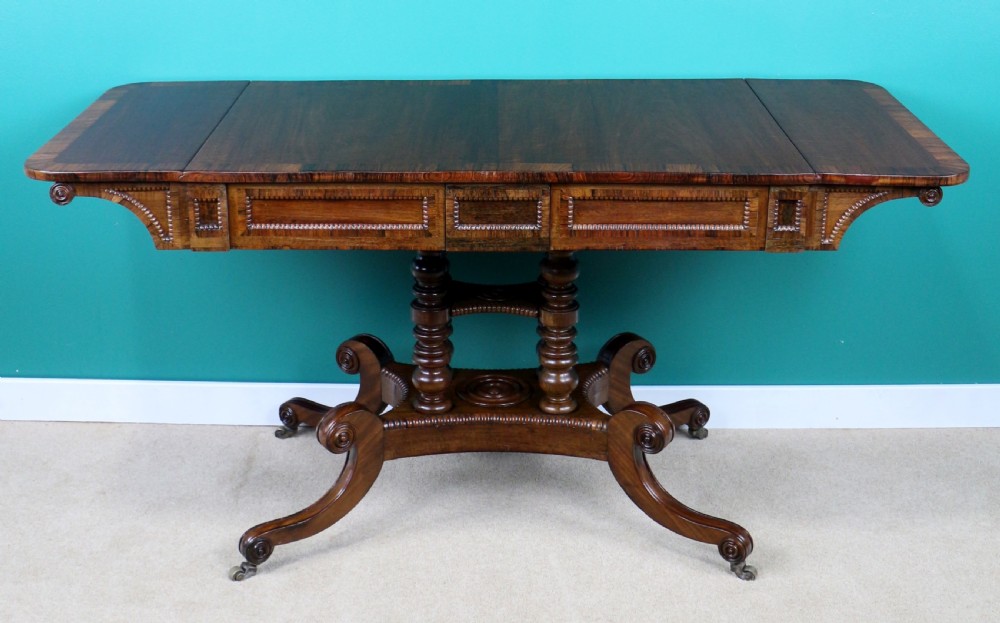 A Regency mahogany and rosewood crossbanded sofa table featuring drop leaves, bead-moulded frieze drawers, turned support pillars, and splayed legs on brass castors. View VI