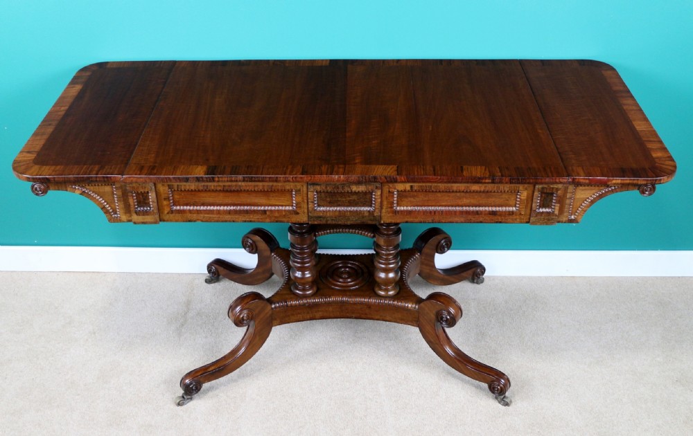 A Regency mahogany and rosewood crossbanded sofa table featuring drop leaves, bead-moulded frieze drawers, turned support pillars, and splayed legs on brass castors. View VII