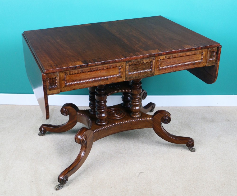 A Regency mahogany and rosewood crossbanded sofa table featuring drop leaves, bead-moulded frieze drawers, turned support pillars, and splayed legs on brass castors. View VIII
