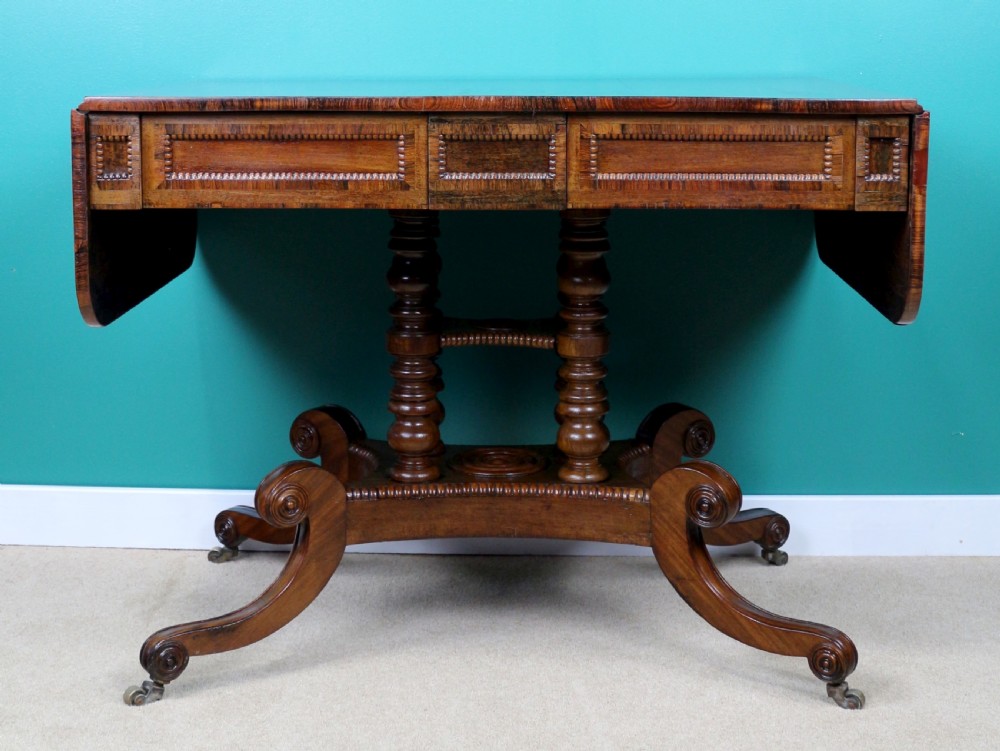 A Regency mahogany and rosewood crossbanded sofa table featuring drop leaves, bead-moulded frieze drawers, turned support pillars, and splayed legs on brass castors. View IX